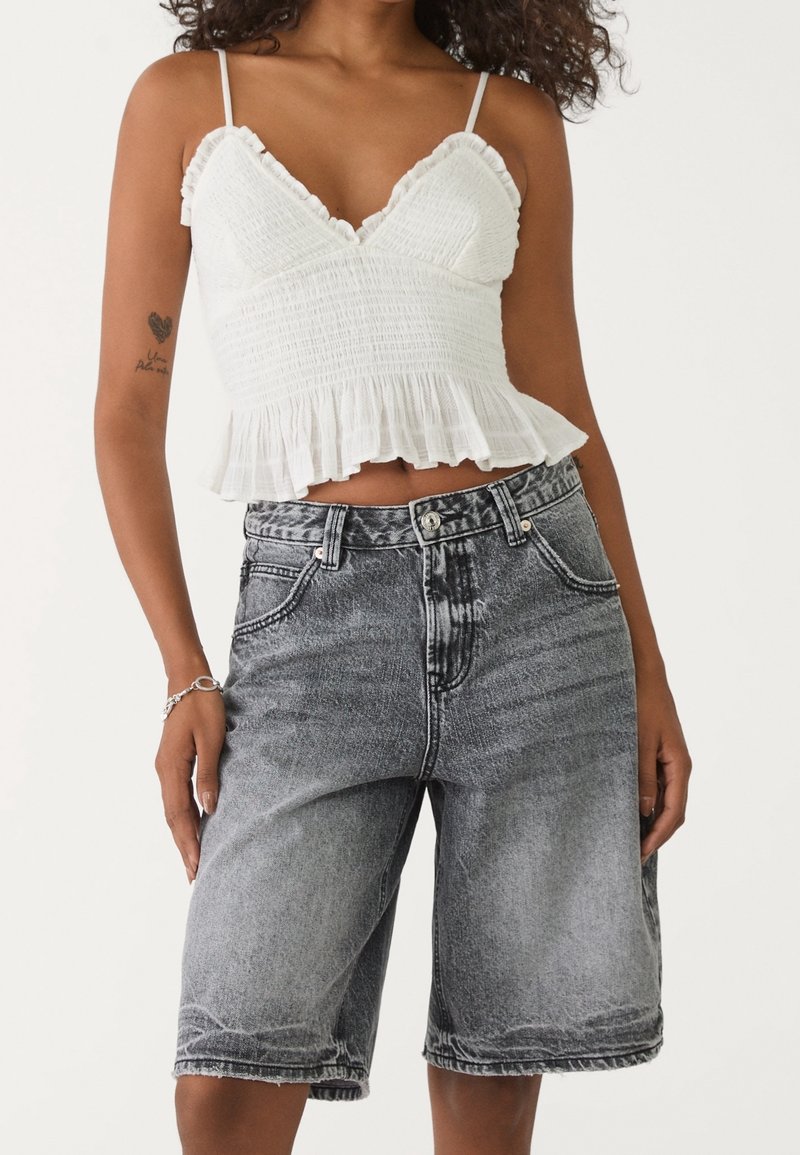 Femme portant un débardeur court blanc à volants et un short en jean noir délavé taille haute jusqu'aux genoux.