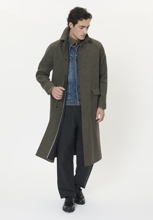 Langer, karierter Mantel in Oliv und Braun, mit hohem Kragen, Frontknöpfen und Seitentaschen, über einer Jeansjacke getragen.