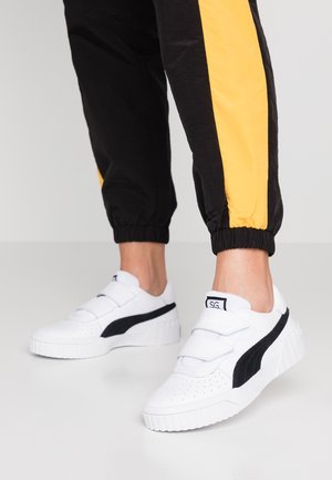 Person iført sorte bukser med gule striber og hvide sneakers med sorte detaljer og velcrolukninger står på et lyst gulv.