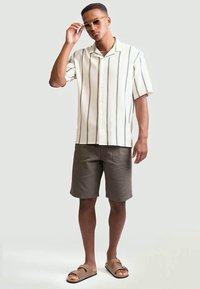 Uomo che aggiusta gli occhiali da sole, indossando una camicia bianca a maniche corte con strisce verticali, pantaloni corti scuri e sandali color sabbia, in piedi su uno sfondo semplice.