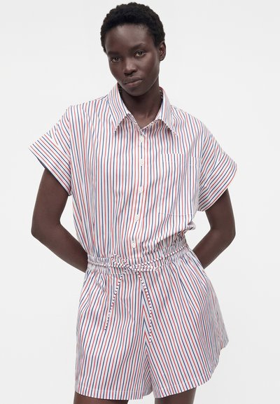 Γυναίκα που φοράει λευκό ολόσωμο σορτς με κόκκινες και μπλε κατακόρυφες ρίγες, κουμπωτό μπροστά και δεμένη ζώνη στη μέση.