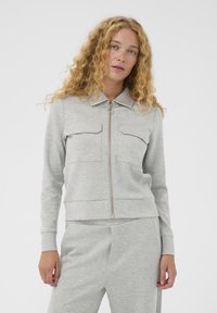 Žena s kučeravými blond vlasmi, oblečená v svetlosivej zipsovej bunde s vreckami na hrudi a zodpovedajúcich nohaviciach na bielom pozadí.