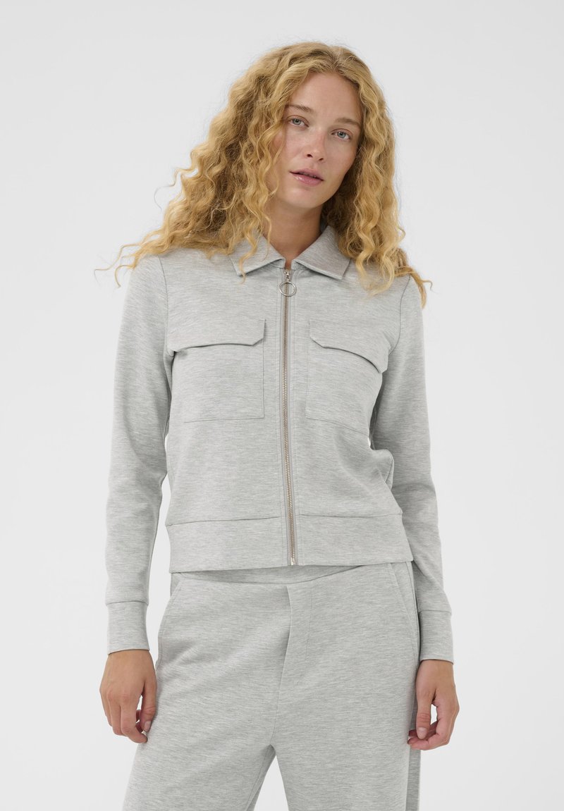 Žena s kučeravými blond vlasmi, oblečená v svetlosivej zipsovej bunde s vreckami na hrudi a zodpovedajúcich nohaviciach na bielom pozadí.