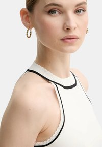 Top bianco a coste senza maniche con scollatura ondulata e bordi neri. Texture liscia, design aderente e estetica minimalista. Orecchini a cerchio dorati.
