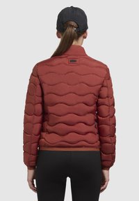 Rode doorgestikte gewatteerde jas met een hoge kraag en elastische manchetten, met een gladde textuur en een strakke uitstraling. Zwarte pet gedragen met een paardenstaart.