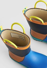 Bottes en caoutchouc jusqu'à la cheville avec des sections bleues, orange et marine. Présentent des poignées en plastique jaune et une doublure douce beige. Semelle texturée pour une meilleure adhérence.