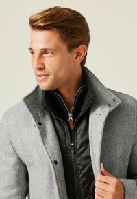 Cappotto grigio a spina di pesce con colletto alto, indossato sopra una giacca nera trapuntata. Presenta bottoni a pressione e una cerniera. Texture liscia.