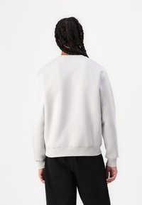 Sweatshirt gris clair avec des poignets et un ourlet côtelés, présentant un design simple et une texture douce, porté par-dessus un pantalon sombre.