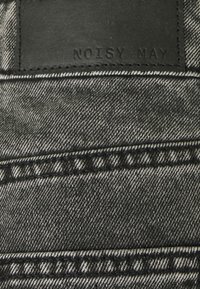 Tela de mezclilla negra con una superficie texturizada, que presenta costuras en contraste, herrajes metálicos y una etiqueta de cuero grabada con "NOISY MAY."