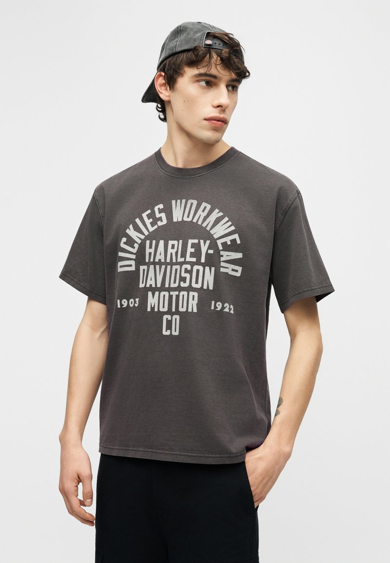 Giovane uomo con cappellino grigio indossato al contrario e t-shirt Harley-Davidson Dickies Workwear grigio scuro, in piedi con una mano in tasca.