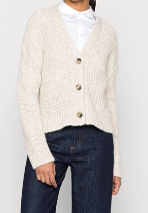 Mujer con cárdigan de punto crema con tres botones sobre una camisa blanca de cuello y jeans de mezclilla azul oscuro, vista desde el cuello hasta las caderas.