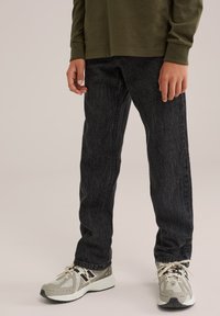 Person som bär svarta jeans och beigea löparskor stående mot en neutral bakgrund.