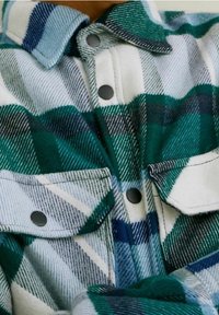 Zielona i biała flanelowa koszula w kratę z wyraźnymi kieszeniami, ciemnymi guzikami i miękką, fakturowaną tkaniną. Kołnierzyk z kontrastowymi paskami.