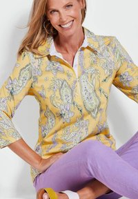 Geel blouse met bloemenpatroon, kraag met witte halslijn, driekwart mouwen, gecombineerd met paarse broek en sandalen met een gele accenten.