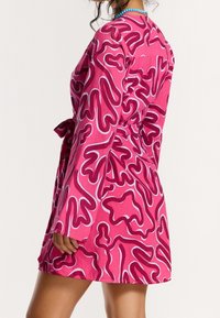 Femme portant une robe rose avec des motifs abstraits tourbillonnants rouges et blancs, une ceinture à la taille et un collier de perles bleu, vue de profil et de dos.