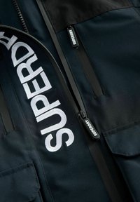 Svart jacka med stort vitt broderat "SUPERDRY"-logotyp, dragkedjefickor och matt yta, med en blandad tygdesign.