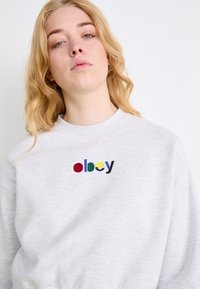 Sudadera gris claro con cuello redondo, que presenta un logo bordado "obey" multicolor en el pecho. Tejido suave y texturizado.