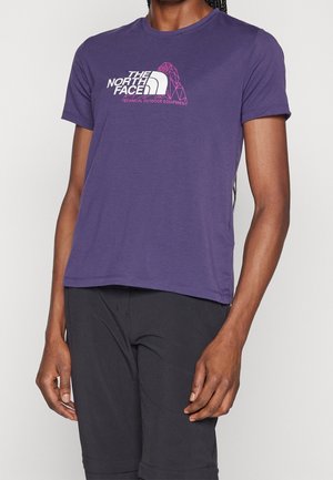 Woman wearing a purple The North Face t-shirt with logo and black outdoor shorts, standing against a plain light background.