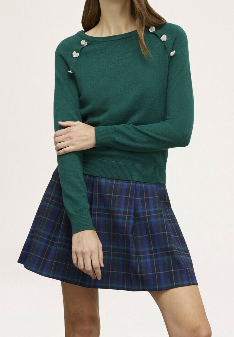 Femme portant un pull vert à manches longues avec des boutons en forme de cœur sur les épaules et une jupe plissée à carreaux bleu et vert.