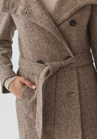 Bruine tweedjas met een dubbelzijdig ontwerp, een ceintuur in de taille, grote revers en een gestructureerde stof met kleurrijke spikkels en zijzakken.