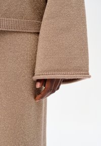 Sweter dziany w miękkim beżu z subtelnymi metalicznymi włóknami. Charakteryzuje się luźnym krojem, szerokimi rękawami oraz ściągaczami na mankietach, podkreślając teksturę i ciepło.