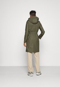 Person iført en olivengrøn bæltejakke med hætte, beige stribede bukser og hvid-grønne sneakers, der står foran en ensfarvet lys baggrund.