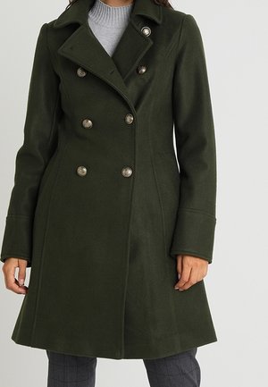 Woman wearing a dark green double-breasted wool coat with silver buttons over a light gray turtleneck sweater and gray checkered pants.