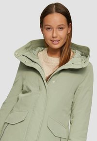 Manteau isolant vert clair avec capuche, doublure matelassée, poches frontales et fermeture éclair. Porté sur un pull en tricot crème.