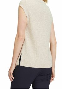 Donna che indossa un maglione a maglia beige senza maniche e pantaloni neri, mostrata di spalle con le braccia rilassate lungo i fianchi.