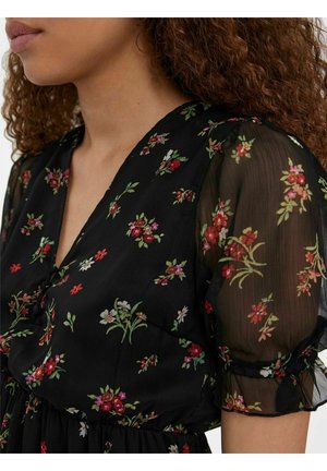 Femme portant une robe noire à fleurs avec des manches courtes bouffantes en voile et un décolleté en V, montrant des cheveux bruns bouclés et le cou.