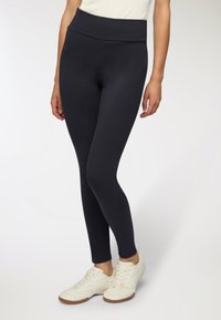 Czarne legginsy o wysokim stanie, wykonane z elastycznej tkaniny, o gładkiej fakturze i dopasowanym kroju, noszone z białymi butami sportowymi.