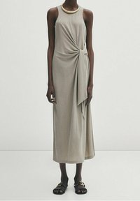 Robe sans manches, longueur genou, en taupe clair avec un détail de nœud tordu à la taille. Tissu fluide, associée à des sandales noires plates.