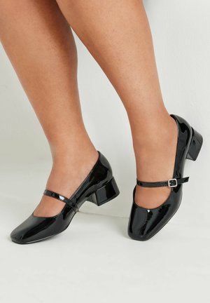 Des chaussures Mary Jane en cuir verni noir avec un bout carré, un petit talon carré et une sangle réglable avec une boucle en argent. Texture lisse et brillante.