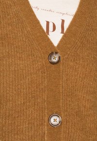 Brauner Cardigan mit V-Ausschnitt, ausgestattet mit einem strukturierten Strickstoff und einem großen, hellen Knopf an der Vorderseite. Die darunterliegende Schicht zeigt bedruckten Text.