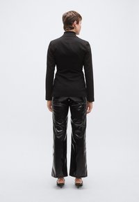 Μαύρο σακάκι ραμμένο στα μέτρα με εφαρμοστή σιλουέτα, συνδυασμένο με γυαλιστερό μαύρο παντελόνι με καμπάνα. Το μοντέλο φαίνεται από πίσω, σε μινιμαλιστικό στυλ.