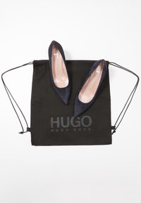 Paire d'escarpins en daim noir à bout pointu posée sur un sac à cordon noir avec le logo "HUGO BOSS" en lettres grises.