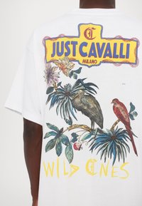Hvit t-skjorte med et fargerikt grafisk motiv som viser tropiske planter, to fugler, samt teksten "JUST CAVALLI MILANO" og "WILD ONES" i gult.