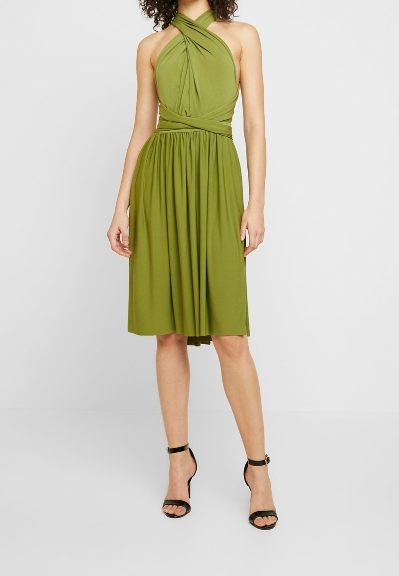 Robe sans manches vert olive avec un col licou torsadé, un corsage ajusté et une jupe plissée, associée à des talons noirs à brides.
