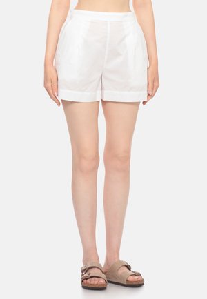 Persona che indossa shorts bianchi a vita alta con tasche laterali e sandali beige, in piedi contro uno sfondo chiaro e semplice, visibile dalla vita ai piedi.