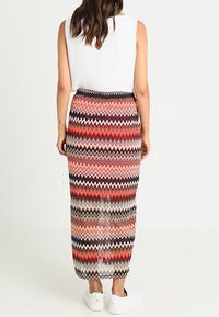 Maxi-Rock mit wellenartigem Muster in den Farben Rot, Pink, Schwarz und Creme. Elastischer Taillenbund. Kombiniert mit einem weißen Oberteil und weißen Sneakers.