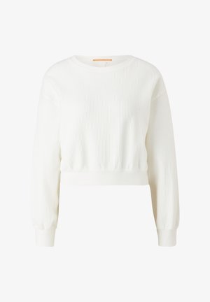 Person trägt weißen, strukturierten, kurz geschnittenen Sweatshirt und passende Jogginghose mit beigen High-Top-Sneakers, Hände in den Taschen.