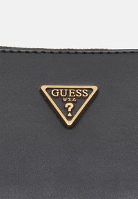 Černý texturovaný materiál se zlatým trojúhelníkovým logem obsahujícím "GUESS USA" a otazník uprostřed, který přidává výrazný akcent.