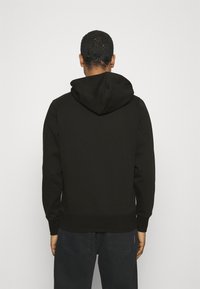 Felpa nera con cappuccio realizzata in tessuto morbido. Presenta una vestibilità ampia, polsini a costine e una tasca a marsupio. Indossata con jeans scuri.