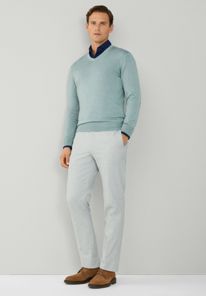 Un pull v-neck bleu clair par-dessus une chemise navy, associé à un pantalon à motifs gris clair et des chaussures en daim marron. Design simple et ajusté.