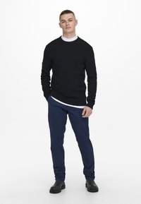 Un pull en laine noir sur un t-shirt blanc à col rond, associé à un pantalon chino bleu marine et des bottes noires. La tenue présente une coupe décontractée et un design simple.