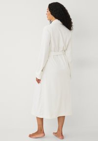 Vrouw met krullend haar, gekleed in een lange witte jurk die op de taille is vastgebonden, staande op blote voeten tegen een effen witte achtergrond, zijwaarts kijkend.
