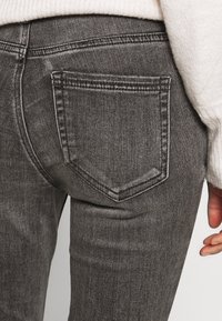 Los jeans de mezclilla gris presentan un bolsillo trasero con un sutil patrón de costura. La tela muestra una textura suave con un aspecto desgastado.