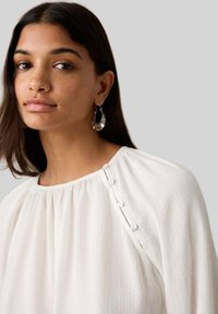 Weiße, strukturierte Bluse mit rundem Ausschnitt und einem Knopfdetail auf einer Schulter. Das Modell trägt silberne Creolen.