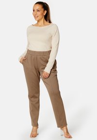 Beige ribbed langemouwtop gecombineerd met bruine taps toelopende broek met zijzakken en een elastische tailleband. Transparante hakken sandalen.