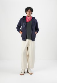 Sudadera navy con cremallera, camiseta de manga corta en verde oscuro, pantalones de pierna ancha color crema y zapatillas blancas con suelas en tono marrón, complementadas con un pañuelo rosa.